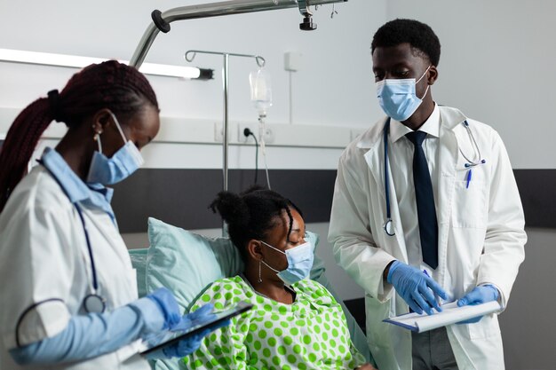 Équipe médicale avec masques faciaux pour prévenir l'infection par le coronavirus surveillant le patient malade lors d'un rendez-vous clinique dans le service hospitalier. médecins afro-américains expliquant les soins de santé