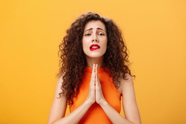 Espoir femme charmante et idiote avec une coupe de cheveux bouclés tenant la main dans la prière mordant la lèvre inférieure dans le besoin ...