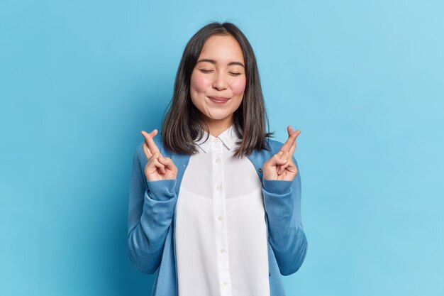 Espoir heureux femme asiatique brune croise les doigts pour la bonne chance prie les rêves deviennent réalité étant bien habillé ferme les yeux