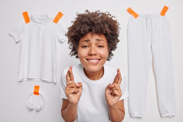 Espoir jeune femme aux cheveux bouclés regarde avec un grand désir garde les doigts croisés mord les lèvres pose à l'intérieur fait voeu isolé sur fond de studio blanc vêtements plâtrés Concept de langage corporel