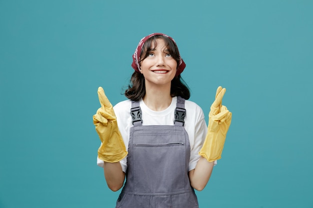 Espoir jeune femme nettoyante portant un bandana uniforme et des gants en caoutchouc levant les yeux tout en mordant la lèvre isolée sur fond bleu