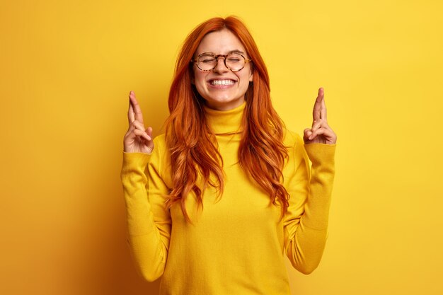 Espoir jeune femme rousse joyeuse sourit ferme largement les yeux garde les doigts croisés croit que les rêves deviennent réalité porte un col roulé décontracté.