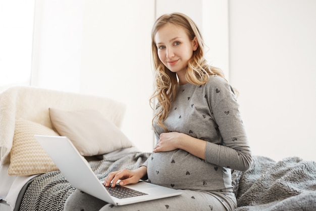 Essayer de créer quelque chose de nouveau pour la future vie de maternité. Gros plan de l'heureuse jeune mère blonde enceinte dans des vêtements gris confortables lisant des articles sur la première année de vie de bébé sur un ordinateur portable.