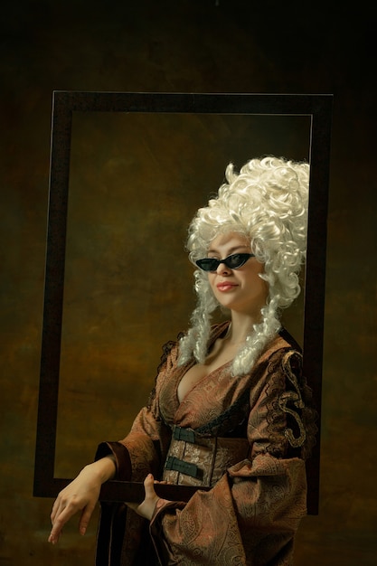 Photo gratuite essayer des lunettes. portrait de jeune femme médiévale en vêtements vintage avec cadre en bois sur fond sombre. modèle féminin en tant que duchesse, personne royale. concept de comparaison des époques, de la mode, de la beauté.
