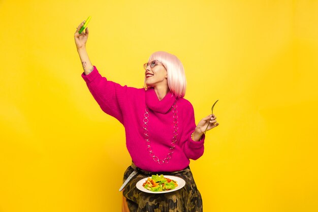 C'est dur d'être influenceur. Selfie d'abord, manger plus tard. Besoin de tirer un plat avant. Portrait de femme caucasienne sur jaune. Beau modèle blonde. Concept d'émotions humaines, expression faciale, ventes, publicité.