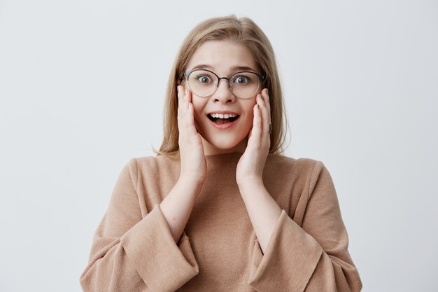 C'est incroyable! Surpris, une femme émue et émue garde les deux mains sur les joues, choquée de voir des prix élevés, isolée contre un mur de studio vierge. Une femme blonde ne peut pas croire aux nouvelles choquantes