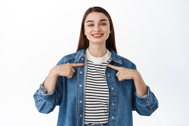 Photo gratuite c'est moi une fille souriante et confiante se pointe du doigt avec un visage fier qui s'auto-prome je suis un geste la présente debout sur fond blanc