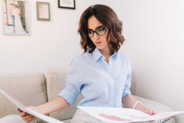 C'est le portrait d'une jeune fille brune assise sur le canapé en studio. Elle porte une chemise bleue et une montre blanche. Elle est curieuse des croquis qu'elle tient à deux mains.