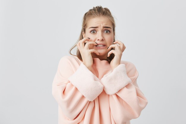 C'est terrible! Femme choquée et effrayée avec des cheveux blonds et raides gardant les deux mains sur les joues et s'exclamant de dégoût et de terreur, les yeux levés, ayant peur en écoutant des histoires d'horreur