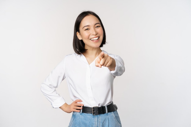 Photo gratuite c'est vous belle jeune femme asiatique chef d'entreprise pointant le doigt vers la caméra et souriant en choisissant d'inviter des personnes recrutant debout sur fond blanc
