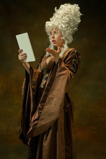 Photo gratuite Étonné à l'aide de la tablette. portrait de jeune femme médiévale en vêtements vintage marron sur fond sombre. modèle féminin en tant que duchesse, personne royale. concept de comparaison des époques, moderne, mode, beauté.
