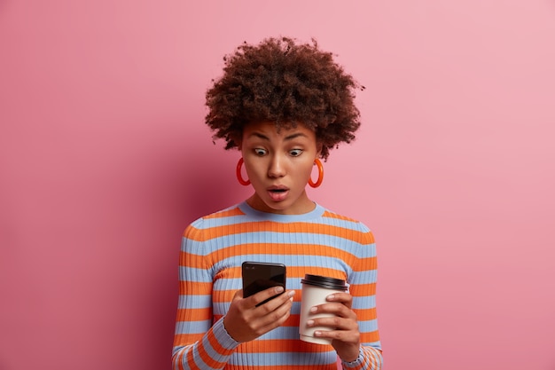 Photo gratuite Étonnée, surprise, une jeune femme bouclée regarde l'écran du téléphone intelligent, voit quelque chose d'étonnant en ligne, lit un message insultant inquiétant, boit du café à emporter, pose contre un mur rose.