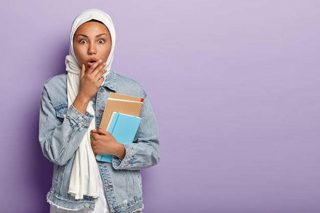 Un étudiant arabe stupéfait fréquente le lycée, a la peau foncée, porte un bloc-notes pour écrire des notes, porte un voile blanc sur la tête, a ses propres traditions religieuses, pose à l'intérieur