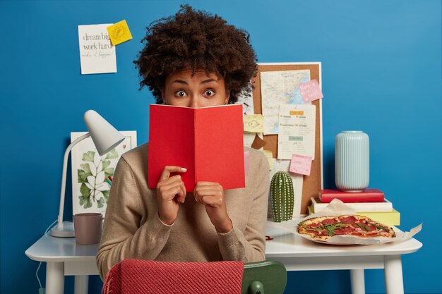 Un étudiant étonné couvre le visage avec un journal rouge, choqué d'oublier une tâche importante à préparer