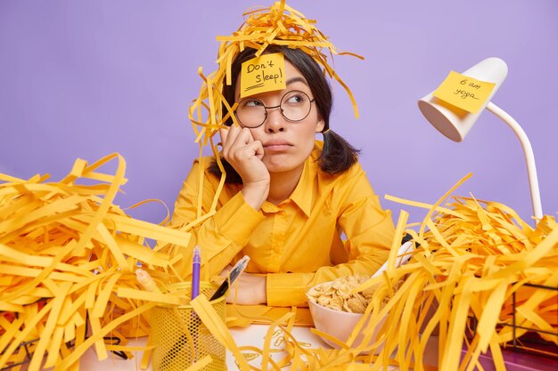 Un étudiant fatigué se prépare pour la session d'examen est assis au bureau avec une expression réfléchie entourée de piles de papier découpé prend des notes sur des autocollants pour ne pas oublier les tâches nécessaires