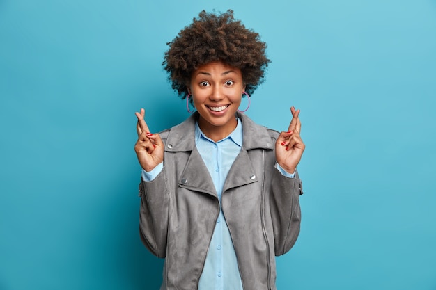 Photo gratuite un étudiant heureux avec une coiffure afro espère réussir l'examen, croise les doigts pour avoir de la chance, fait des vœux, anticipe des résultats positifs, porte des vêtements à la mode, pose