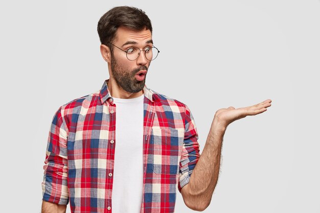 Étudiant homme barbu émotionnel avec expression surprise maintient la paume contre le mur blanc