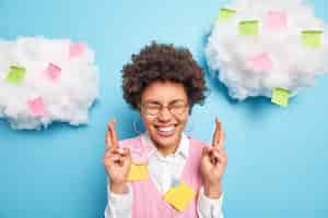 Photo gratuite un étudiant optimiste avec des cheveux bouclés foncés garde les doigts croisés croit en la bonne chance à l'examen porte une chemise blanche et un gilet pose contre le mur bleu