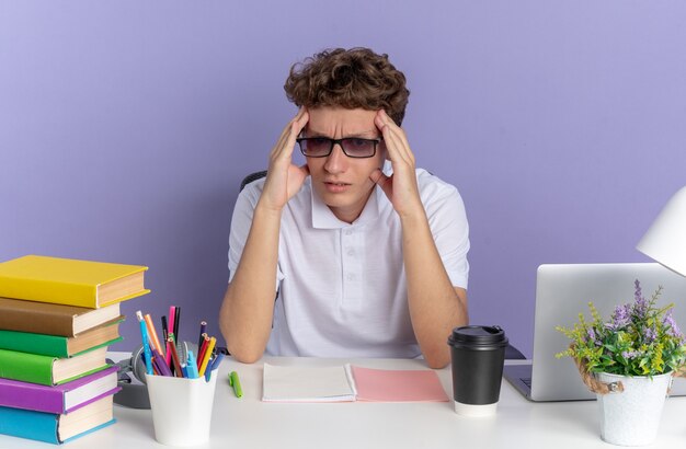 Un étudiant en polo blanc portant des lunettes assis à la table avec des livres ayant l'air fatigué et ennuyé de toucher sa tête sur une surface bleue