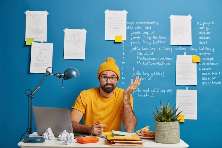 Photo gratuit un étudiant de sexe masculin inquiet et incertain résout un problème difficile tout en travaillant dans une salle d'étude, écrit des informations, fait une dissertation, manque d'idée, porte des vêtements jaunes