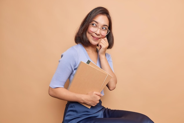 Photo gratuite l'étudiant tient des cahiers à spirale garde la main sur la joue regarde au loin avec une expression rêveuse pense aux vacances et au repos après avoir étudié porte de grandes lunettes optiques jupe pull bleue