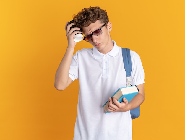 Un étudiant en vêtements décontractés portant des lunettes avec un sac à dos tenant un livre et une tasse en papier ayant l'air fatigué
