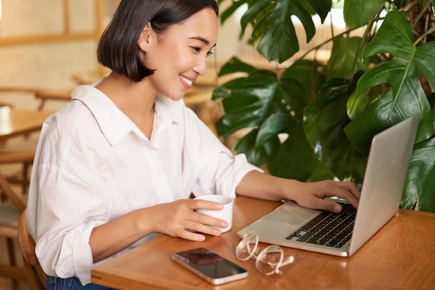 Photo gratuite Étudiante assise dans un café avec une tasse de café, jeune femme asiatique travaillant sur un ordinateur portable dans un restaurant assis