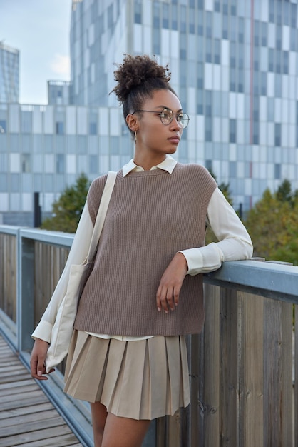 Photo gratuite une étudiante aux cheveux bouclés peignés porte des vêtements soignés concentrés dans des poses à distance au pont contre les gratte-ciel porte un sac en tissu avec les choses nécessaires.