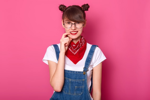 Une étudiante aux cheveux noirs sourit, garde l'index sur sa lèvre, a l'air timide. Jeune fille porte un t-shirt, une salopette en jean avec un bandana rouge sur le cou.