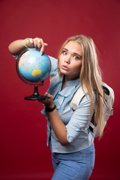 Une étudiante blonde tient un globe et a l'air surprise.