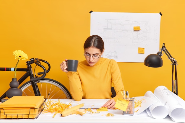 Une étudiante concentrée d'une faculté d'architecture fait ses devoirs réfléchit à des idées créatives boit du café est assise dans un espace de coworking crée des croquis et des plans développe son propre entrepreneuriat social