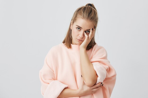 Étudiante Européenne Ennuyée Portant Un Sweat-shirt Rose élégant Touchant Le Visage Avec La Main, L'air Ennuyé, Fatigué D'écouter Des Histoires Sans Intérêt. Le Langage Du Corps