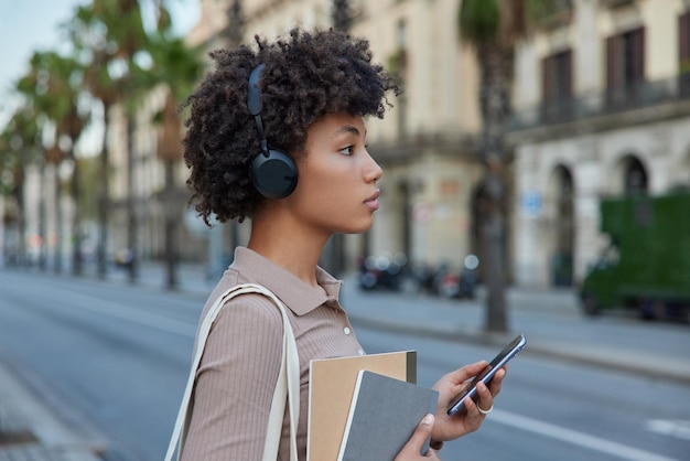 Photo gratuite une étudiante frisée et réfléchie porte des manuels et des blocs-notes contient des recherches de smartphones modernes sur des chansons populaires via une application de média cellulaire pour écouter sur des stands de loisirs sur une route vide en ville