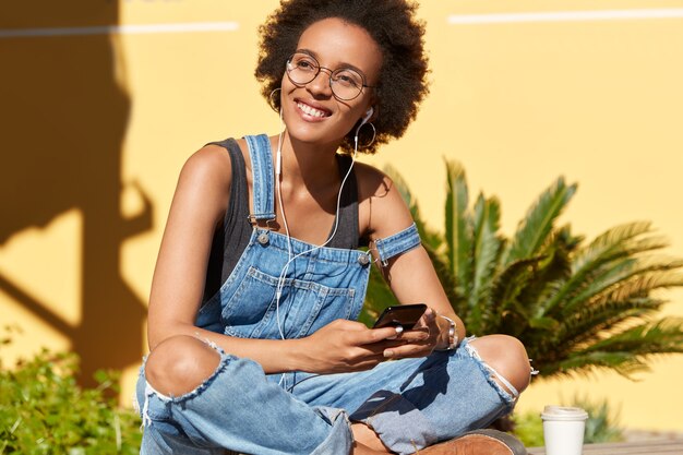 Une étudiante joyeuse écoute un livre audio dans des écouteurs, a des cours en ligne, aime passer du temps dans un endroit tropical, garde les jambes croisées, porte une salopette décontractée en denim, se concentre sur le côté avec un sourire agréable