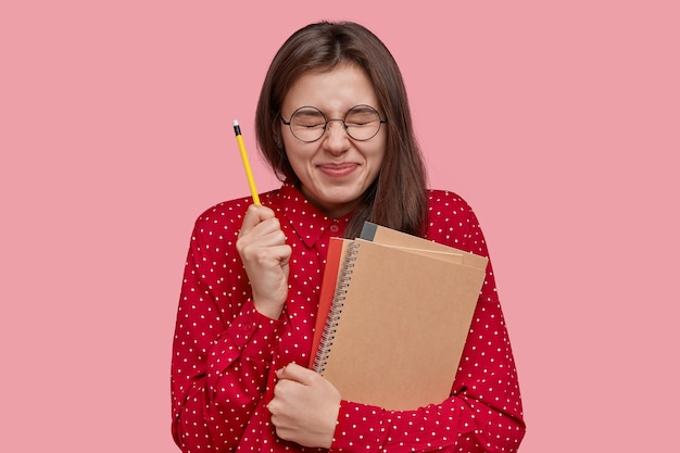 Une étudiante pleine d'espoir ferme les yeux, garde un crayon à la main, porte des blocs-notes, croit entendre le résultat de l'examen, porte des lunettes rondes et un chemisier rouge, isolé sur un mur rose du studio