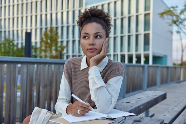 Une étudiante réfléchie écrit des notes dans un cahier tient un stylo a une expression pensive met ses pensées dans son journal personnel pendant les loisirs porte des vêtements soignés fait une liste de contrôle des enregistrements de l'horaire