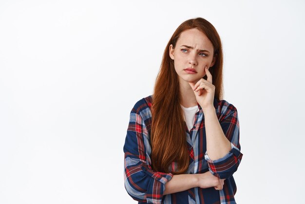 Une étudiante rousse perplexe a l'air réfléchie ou perplexe en fronçant les sourcils en pensant prendre une décision difficile debout sur fond blanc et en réfléchissant