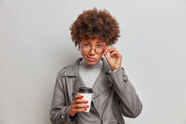 Une étudiante sérieuse regarde avec confiance à travers des lunettes transparentes, écoute attentivement l'intercoluteur, boit du café à emporter, vêtue d'une veste grise