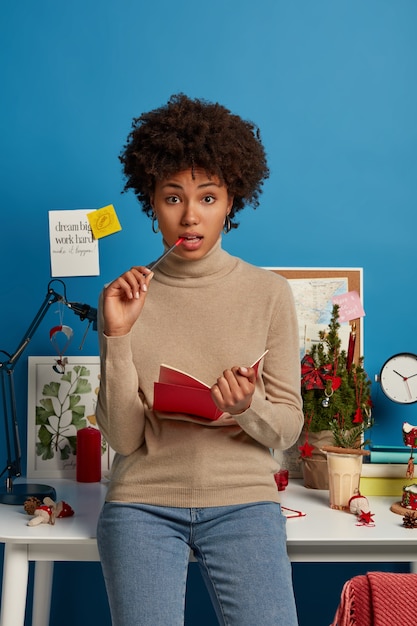 Photo gratuite des étudiantes sérieuses et frisées, songeuses, rédigent un essai dans un manuel, occupent une place d'éducation et d'autodidacte, prennent des notes dans le bloc-notes, posent près du bureau dans la salle d'étude, font leurs devoirs, s'habillent avec désinvolture.