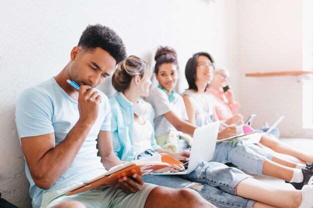 Des étudiants assis par terre avec des ordinateurs portables et faisant leurs devoirs ensemble. Garçon noir avec une coiffure à la mode pensant à une tâche difficile, tenant un stylo à la main.