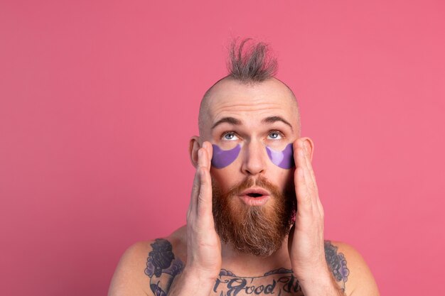 Européen bel homme topless tatoué barbu avec masque de patchs oculaires violet posant à la caméra sur rose