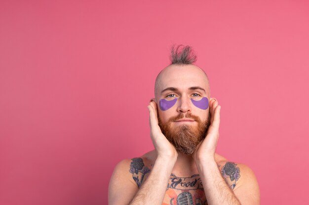 Européen bel homme topless tatoué barbu avec masque de patchs oculaires violet posant à la caméra sur rose