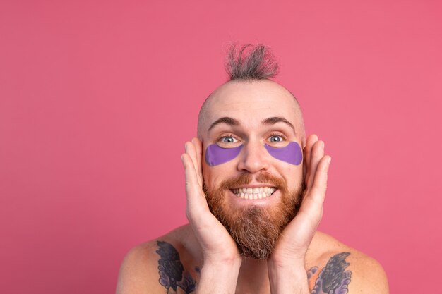 Européen bel homme topless tatoué barbu avec masque de patchs oculaires violet posant à la caméra sur rose