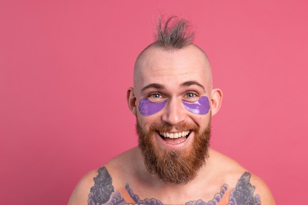 Européen bel homme topless tatoué barbu avec masque de patchs oculaires violet posant à la caméra sur rose