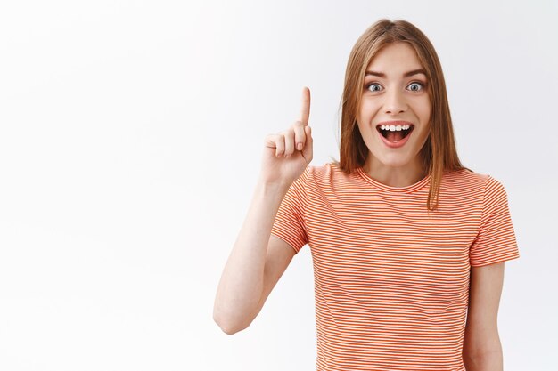 Excellente idée, la fille a trouvé la solution. Belle femme joyeuse et amusée en t-shirt rayé levant un doigt, ampoule, geste eurêka, souriant fasciné, suggère quelque chose de génie, fond blanc