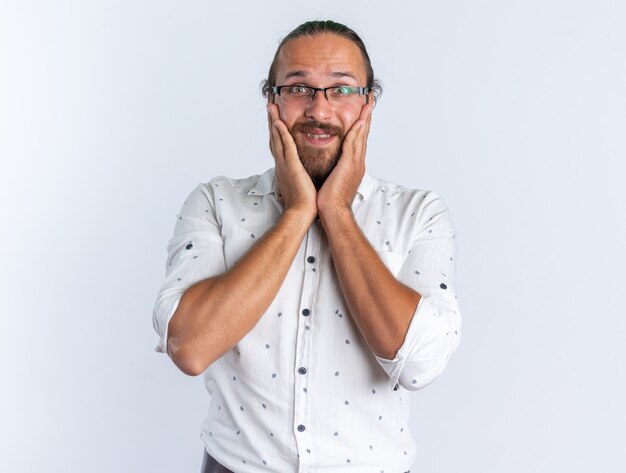 Excité bel homme adulte portant des lunettes en gardant les mains sur le visage en regardant la caméra isolée sur le mur blanc