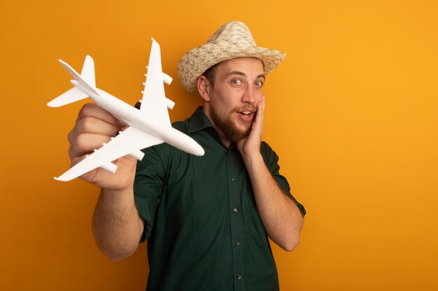 Excité bel homme blond avec chapeau de plage détient avion modèle isolé sur mur orange