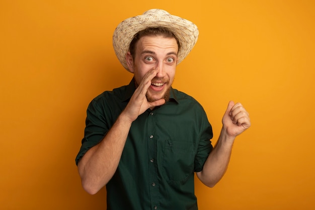 Excité bel homme blond avec chapeau de plage tient la main près de la bouche et garde le poing isolé sur le mur orange