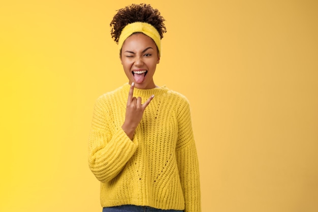 Excité charmante souriante audacieuse jeune femme afro-américaine s'amusant à montrer la langue rock-n-roll heavy metal geste clignant joyeusement en profitant d'une fête géniale, debout sur fond jaune.