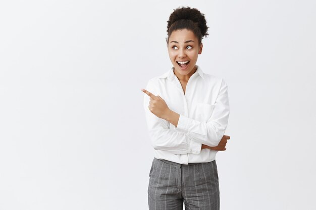 Excité femme afro-américaine en chemise blanche, regardant et pointant en haut à gauche étonné